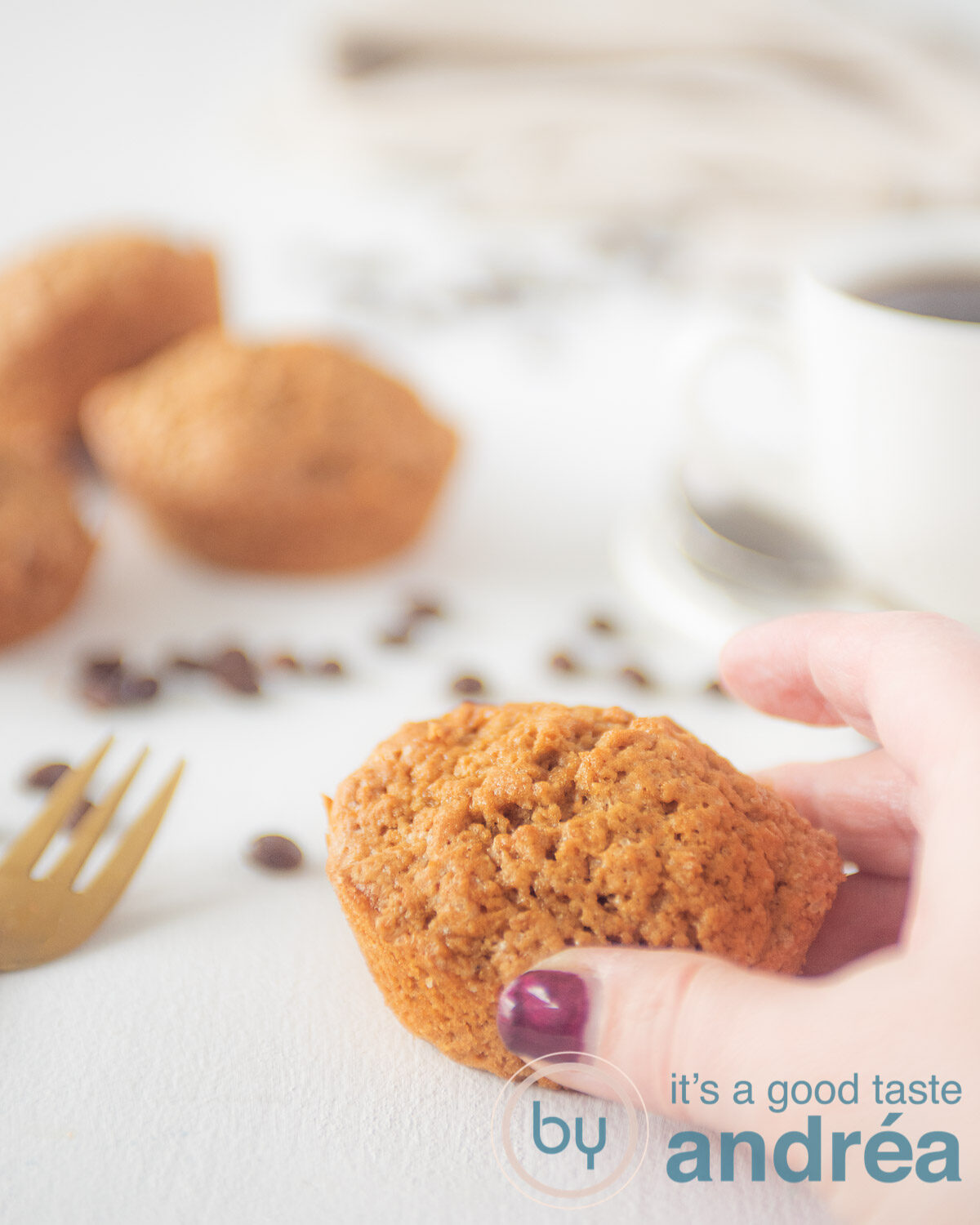 Een hand die een muffin van de tafel pakt. Een kopje koffie en extra muffins in de achtergrond.