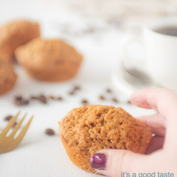 Een hand die een muffin van de tafel pakt. Een kopje koffie en extra muffins in de achtergrond.