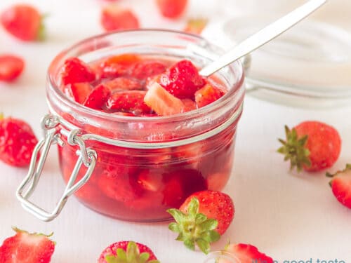 Een vierkante foto met een glazen pot gevuld met aardbeien. Er omheen verspreid aardbeien. Een lepel steekt erin.