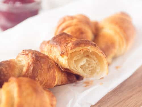 Een vierkante foto met een bakpapier op een houten plank met vier croissants en een halve croissant waar je de luchtige binnenkant goed kunt zien.