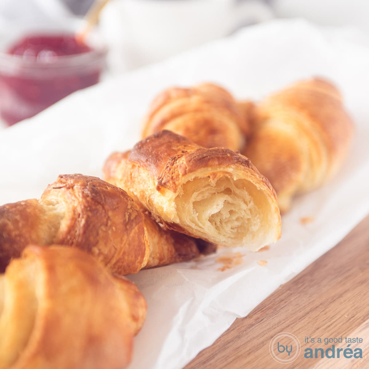 Een vierkante foto met een bakpapier op een houten plank met vier croissants en een halve croissant waar je de luchtige binnenkant goed kunt zien.