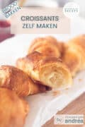 Een aantal croissants op een wit bakpapier op een houten plank. Een tekst bovenin: croissants zelf maken, brood bakken, makkelijk recept.