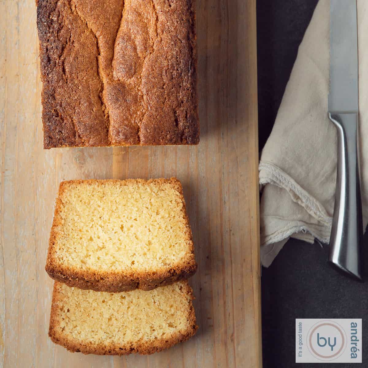 Een vierkante foto met een deel van een houten plank met een cake waar twee plakjes afgesneden zijn en ervoor liggen. Aan de rechterkant ligt een mes.