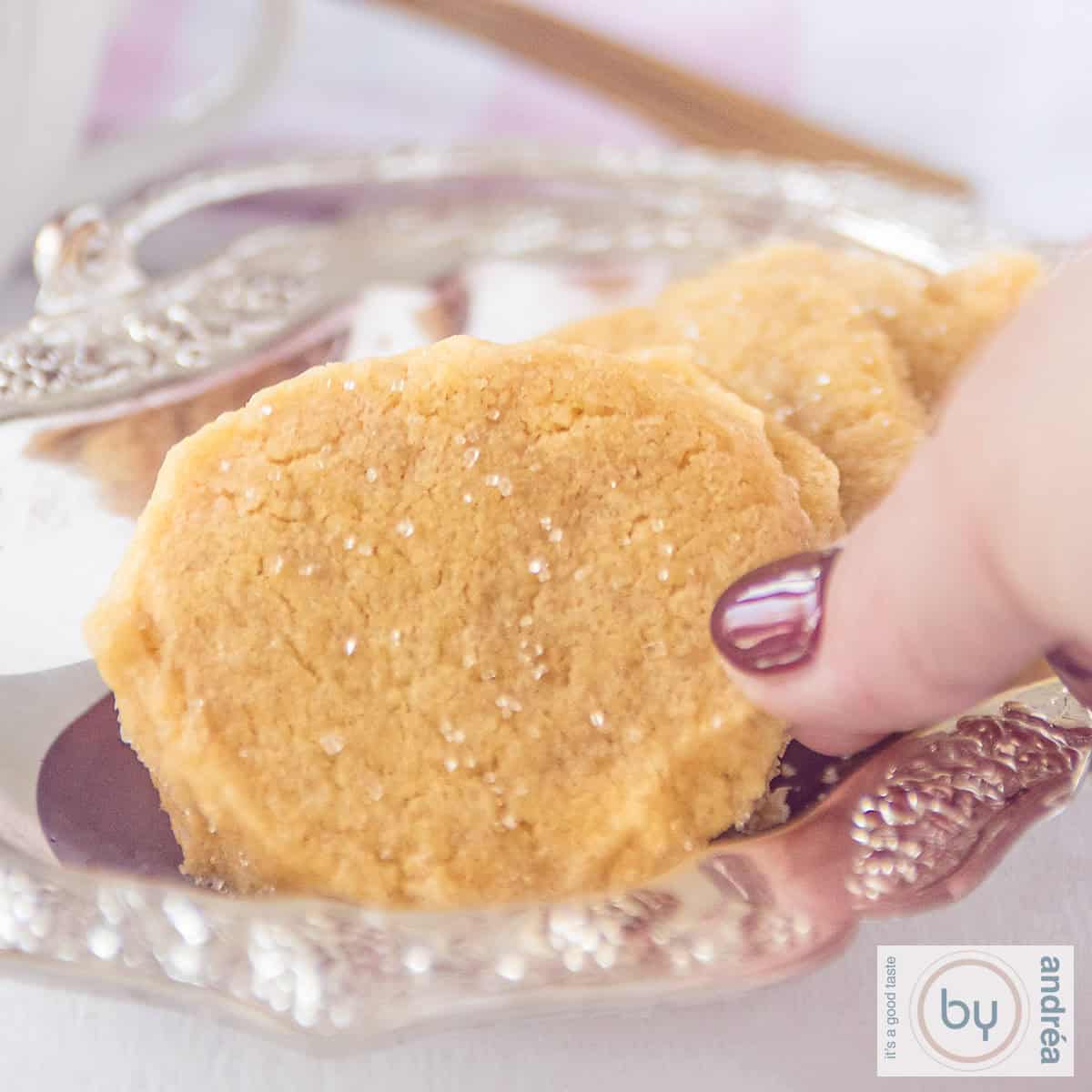 Een vierkante foto met een deel van een zilveren schaal met koekje met een vleugje gember. Een hand pakt een koekje.