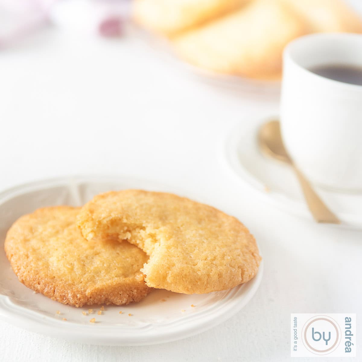 Een vierkante foto met een wit bord met twee koekjes, van een een hap gebeten op een witte ondergrond. Een kop koffie in de achtergrond