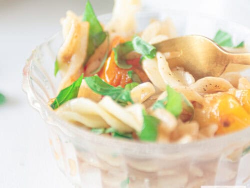 Een vierkante foto met een glazen schaaltje met een pastasalade met gestoofde tomaatjes
