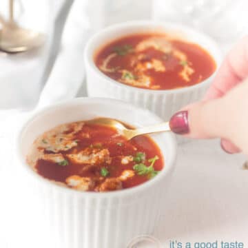 Twee witte kopjes gevuld met tomatensoep, gehaktballetjes en een drupje room. Een hand die een lepel vasthoud die een hapje neemt.