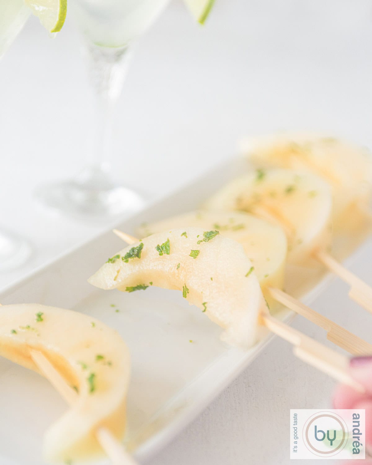 Een witte schaal met vijf gemarineerde galia meloenpartjes in mojito marinade op een rij. Een hand pakt een partje.