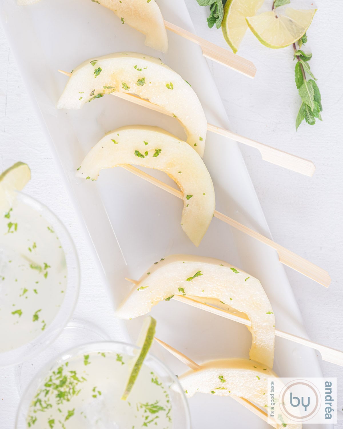 Een foto van bovenaf van een witte schaal met mojito meloen lolly's op een rij. Twee cocktails onderin. Bovenin wat garnering.