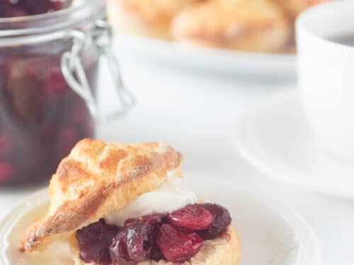 scones met cranberry limoen compote en clothed cream op een wit bord op een witte ondergrond. In de achtergrond een glazen pot met cranberry, een bord met scones en een kopje thee.