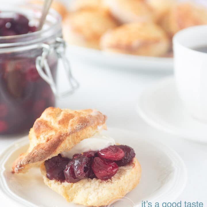scones met cranberry limoen compote en clothed cream op een wit bord op een witte ondergrond. In de achtergrond een glazen pot met cranberry, een bord met scones en een kopje thee.
