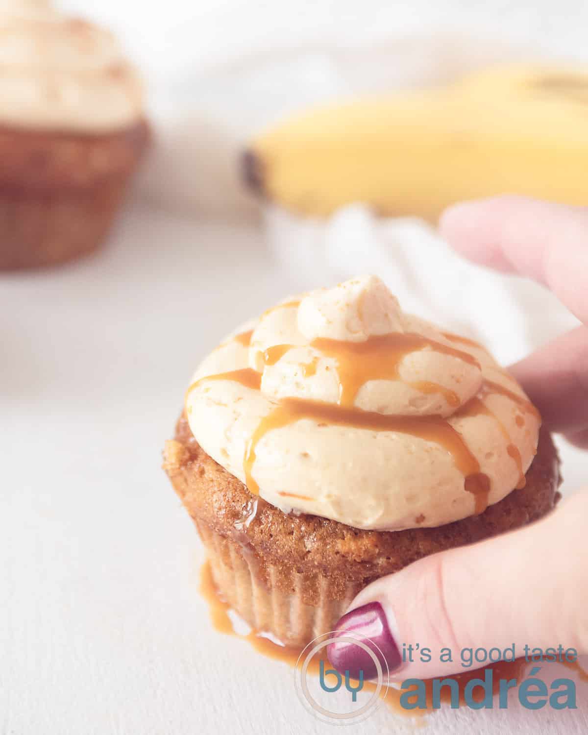 Een bananencupcake met karamel creme topping. Een hand pakt de cupcake. Banaan en een andere cupcake in de achtergrond