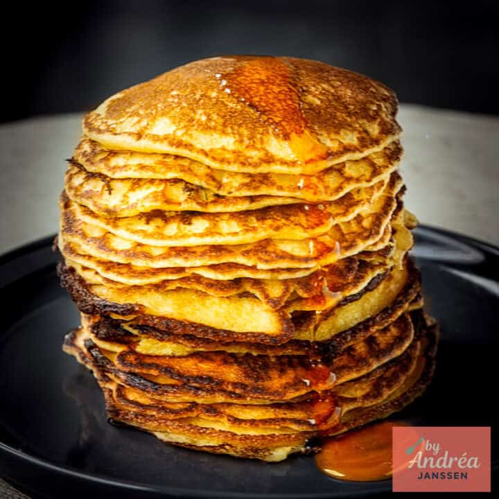 A square picture with a black plate a pile of buttermilk cornflour pancakes and some maple syrup drizzled on top.