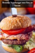 Een hamburger met broodje, tomaat, bacon en sla en gevuld met feta. Een tekst bovenin: broodje hamburger met feta en bacon