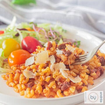 Een vierkante foto van een wit bord met macaroni gevuld met ui, gehakt, spekjes en tomatenpuree. Een salade ernaast.