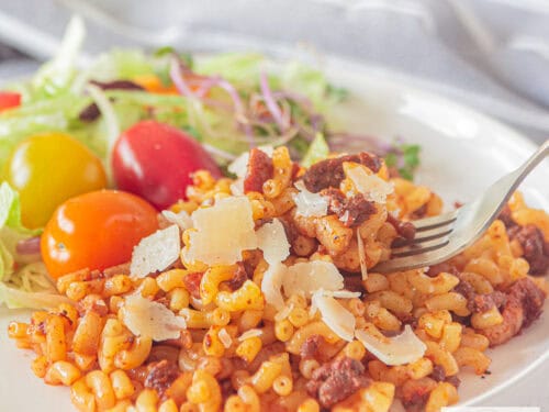 Een vierkante foto van een wit bord met macaroni gevuld met ui, gehakt, spekjes en tomatenpuree. Een salade ernaast.