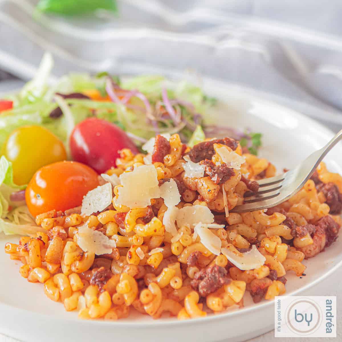 Een vierkante foto van een wit bord met macaroni gevuld met ui, gehakt, spekjes en tomatenpuree. Een salade ernaast.
