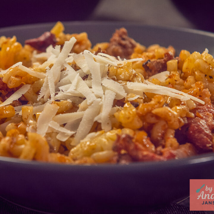 Macaroni met gehakt en spekjes in een donker bord, met geschaafde Parmezaanse kaas erop.