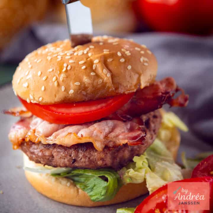 Een vierkante foto met een burger met feta gevuld op een broodje met tomaat, bacon, sla.