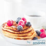 Een vierkante foto van een stapel pancakes met vers fruit op een wit bord.