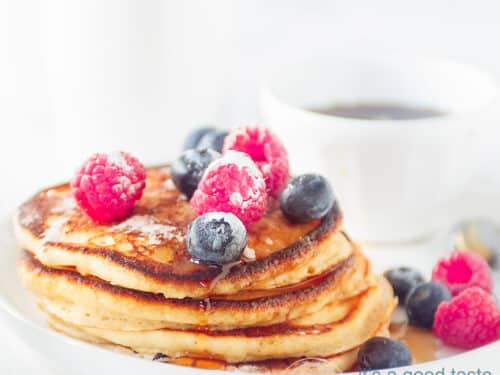 Een vierkante foto van een stapel pancakes met vers fruit op een wit bord.