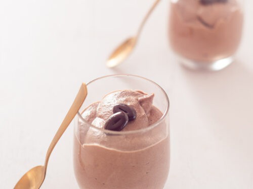 Een vierkante foto met twee glaasjes gevuld met chocolademousse met twee gouden lepels die tegen het glas staan