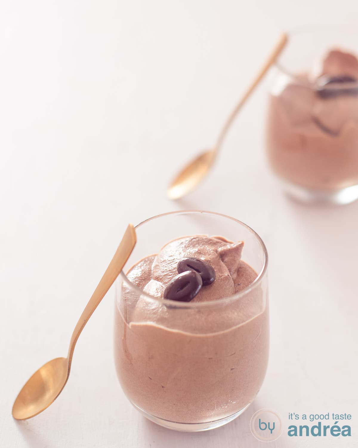 A height photo with two glasses filled with milk chocolate mousse. A gold spoon rests against the glasses. A white background.
