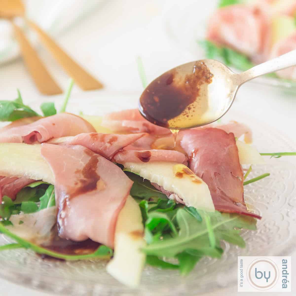 Een vierkante foto met een bord met rucola, pancetta en schijven meloen. Dressing wordt er op gedruppeld!