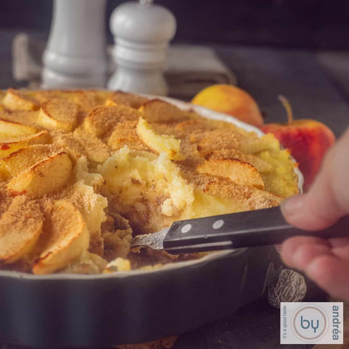 Een vierkante foto van een deel van een ovenschotel met stoofvlees, aardappelpuree, appeltjes. Een schep eten wordt eruit genomen.