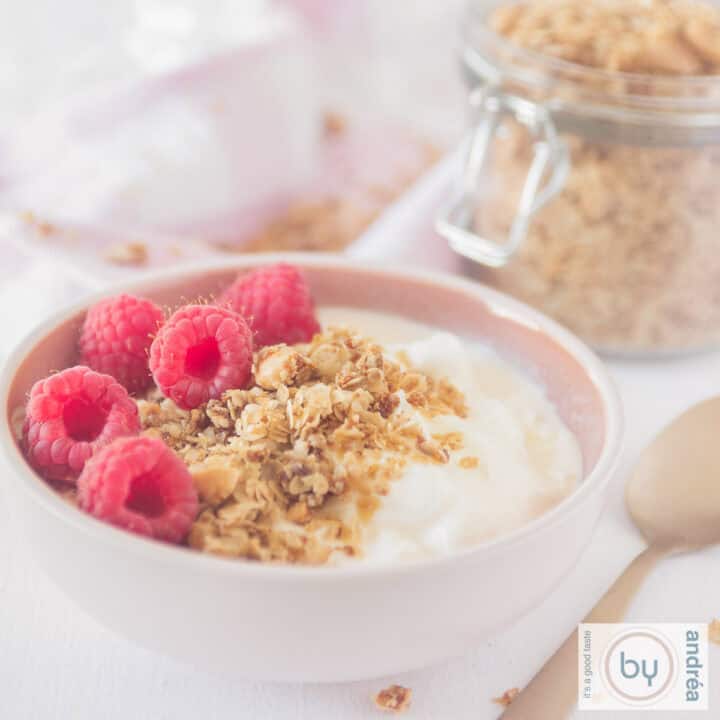 Een vierkante foto met een roze kom gevuld met yoghurt, frambozen en amandelgranola. Nog wat granola op de achtergrond.