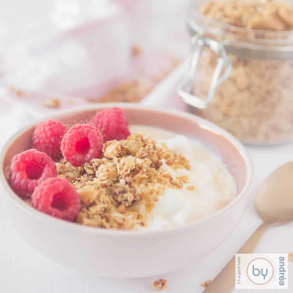 Een vierkante foto met een roze kom gevuld met yoghurt, frambozen en amandelgranola. Nog wat granola op de achtergrond.