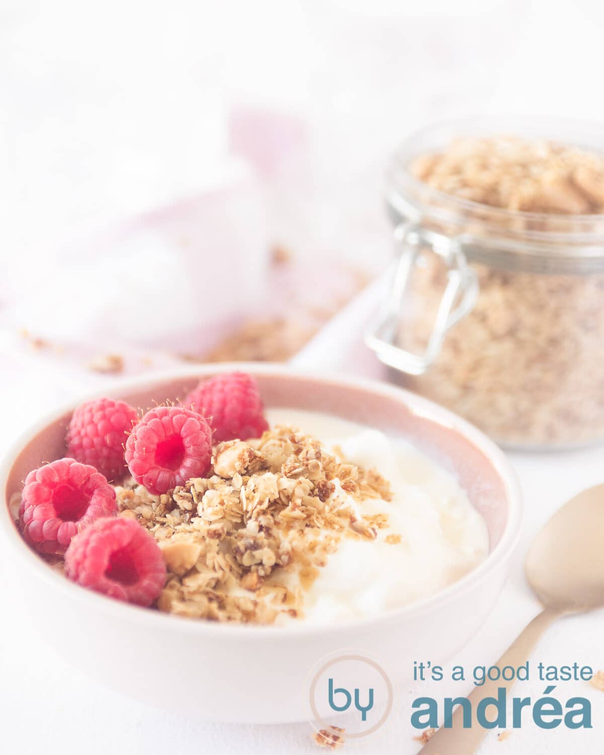 een roze bowl gevuld met yoghurt, granola en frambozen. Rechtsachter een potje met granola. een gouden lepel aan de rechterkant.