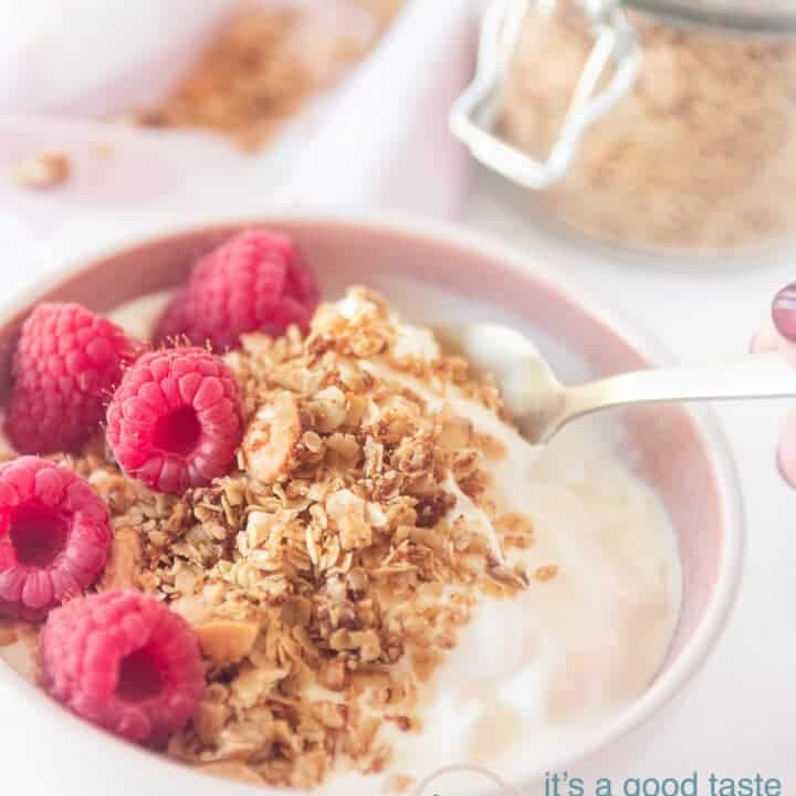 Een roze schaaltje gevuld met cruesli, frambozen en yoghurt. Een pot met cruesli in de achtergrond