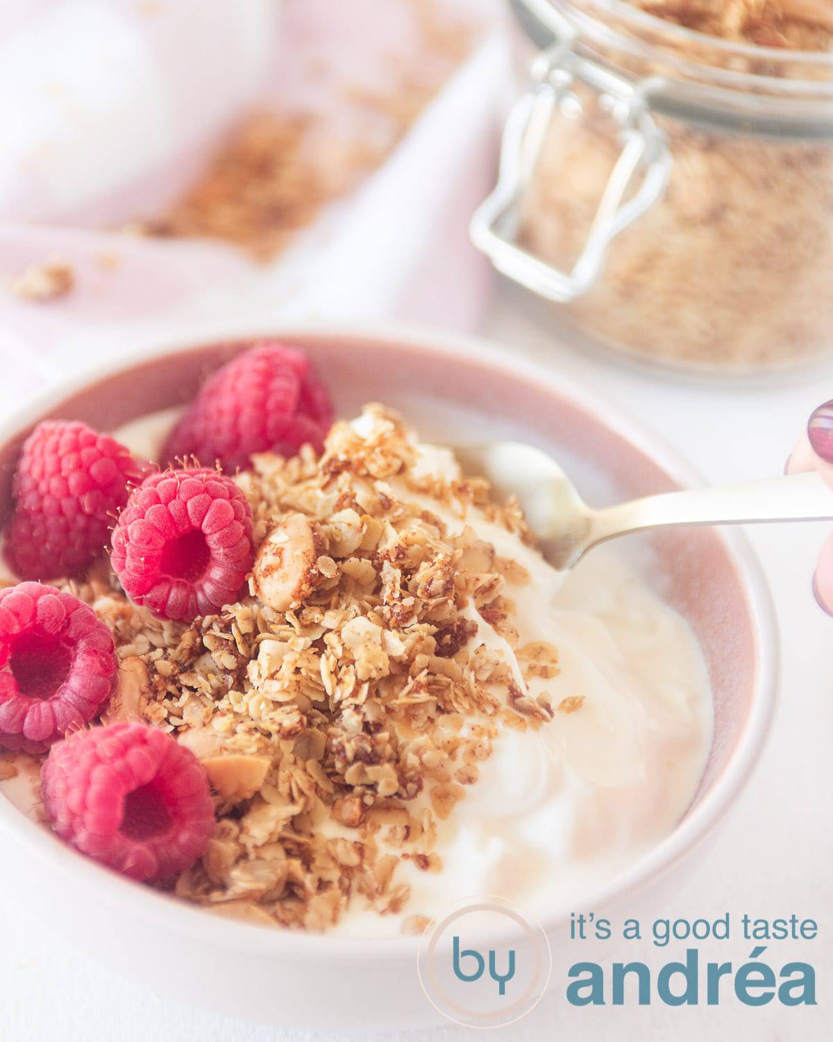 Een roze schaaltje gevuld met cruesli, frambozen en yoghurt. Een pot met cruesli in de achtergrond