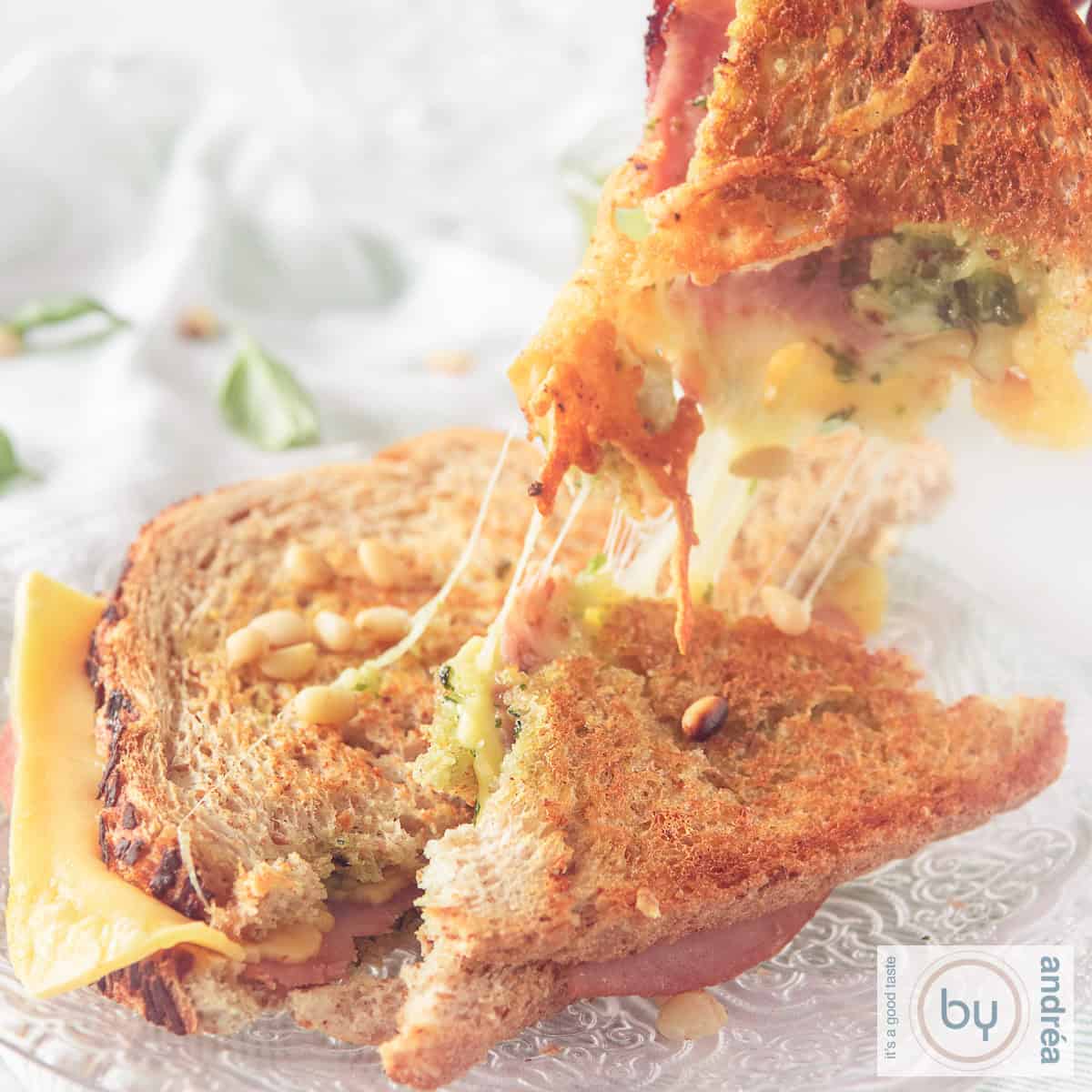 Een vierkante foto met een tosti, gehalveerd. Een half stuk is losgebroken en wordt omhoog gehouden. Gesmolten kaas slierten tussen de stukken.