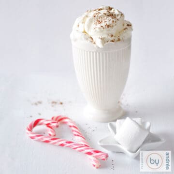Een vierkante foto met een witte mok gevuld met witte chocolademelk. In e voorgrond marhsmallows en kerststokjes