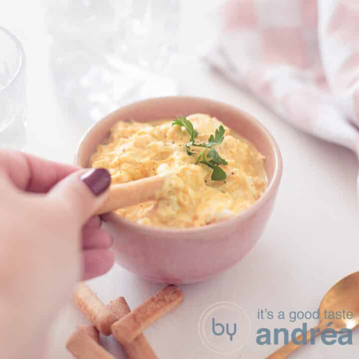 Een vierkante foto van een roze bakje gevuld met een eiersalade zonder mayonaise. Een tapas stokje schept er wat uit. Een witte ondergrond. Rechtsbovenin een roze witte theedoek