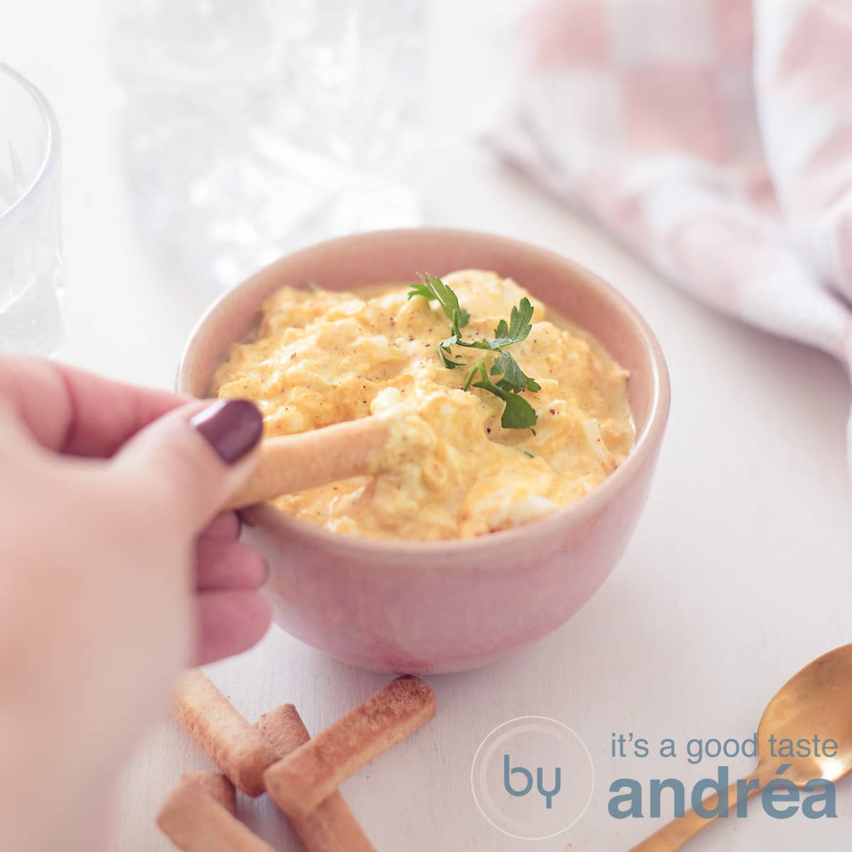 Een vierkante foto van een roze bakje gevuld met een eiersalade zonder mayonaise. Een tapas stokje schept er wat uit. Een witte ondergrond. Rechtsbovenin een roze witte theedoek