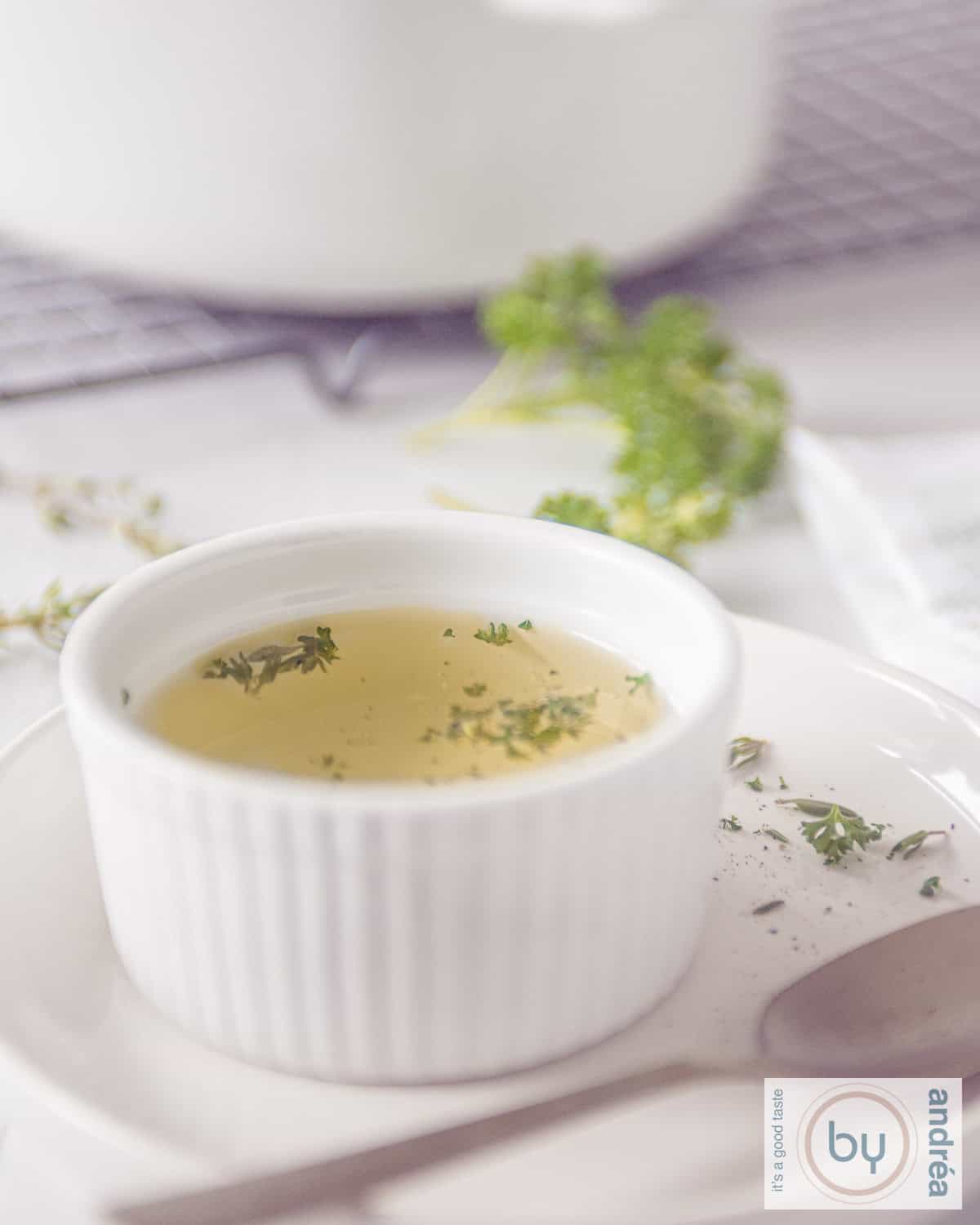 Een witte kom gevuld met groentebouillon. Kruiden eromheen en in de achtergrond de soeppan.