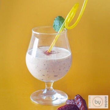 Een vierkante foto met een gele achtergrond. In de voorgrond een mango kokos smoothie met dadels.