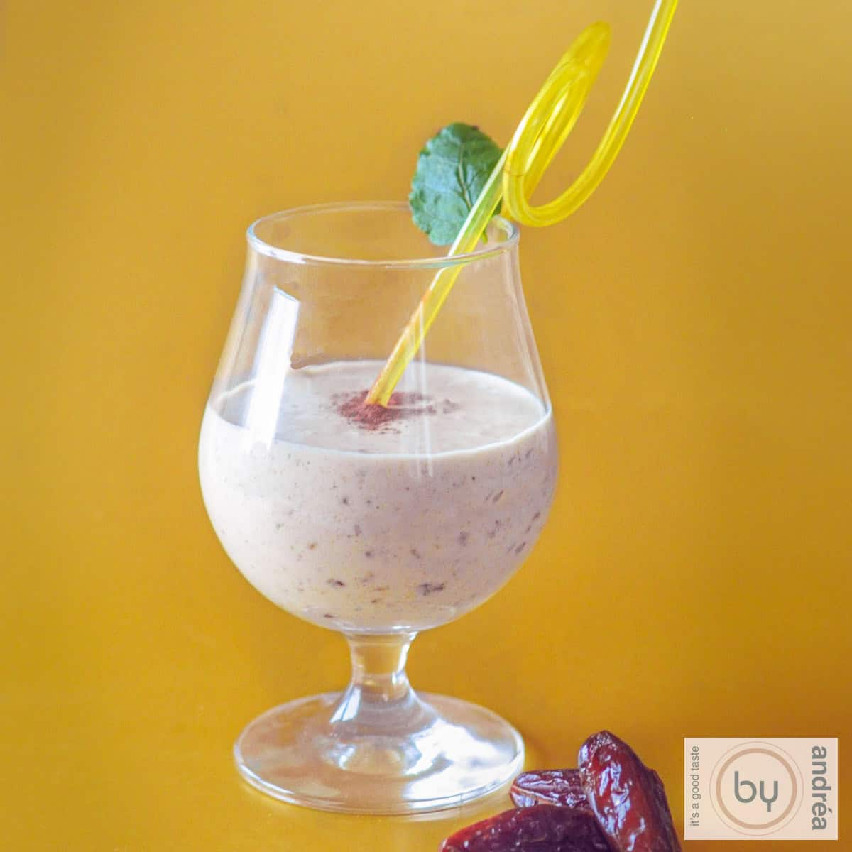 Een vierkante foto met een gele achtergrond. In de voorgrond een mango kokos smoothie met dadels.