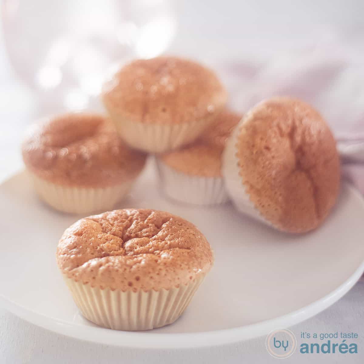 Een vierkante foto met een wit bord met vijf zoete melk cupcakes, een in de voorgrond. Een witte ondergrond.