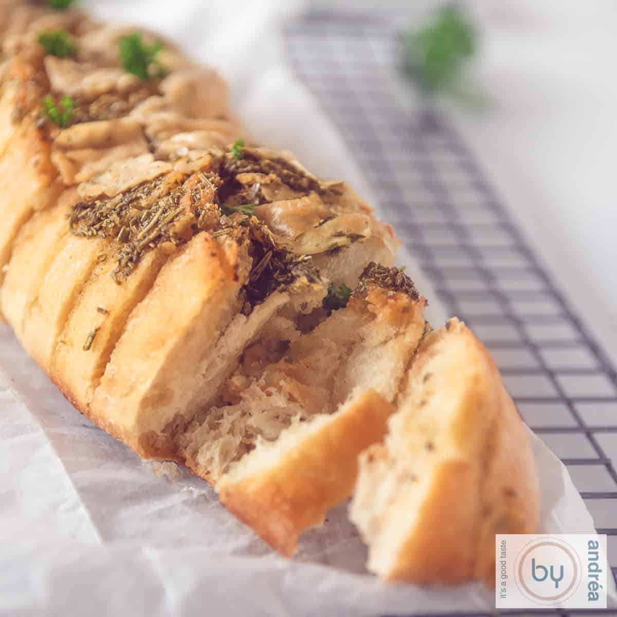 Een vierkante foto met een stokbrood op een bakpapier, waar twee plakjes af zijn gesneden en de knoflookolie en kruiden zichtbaar zijn.