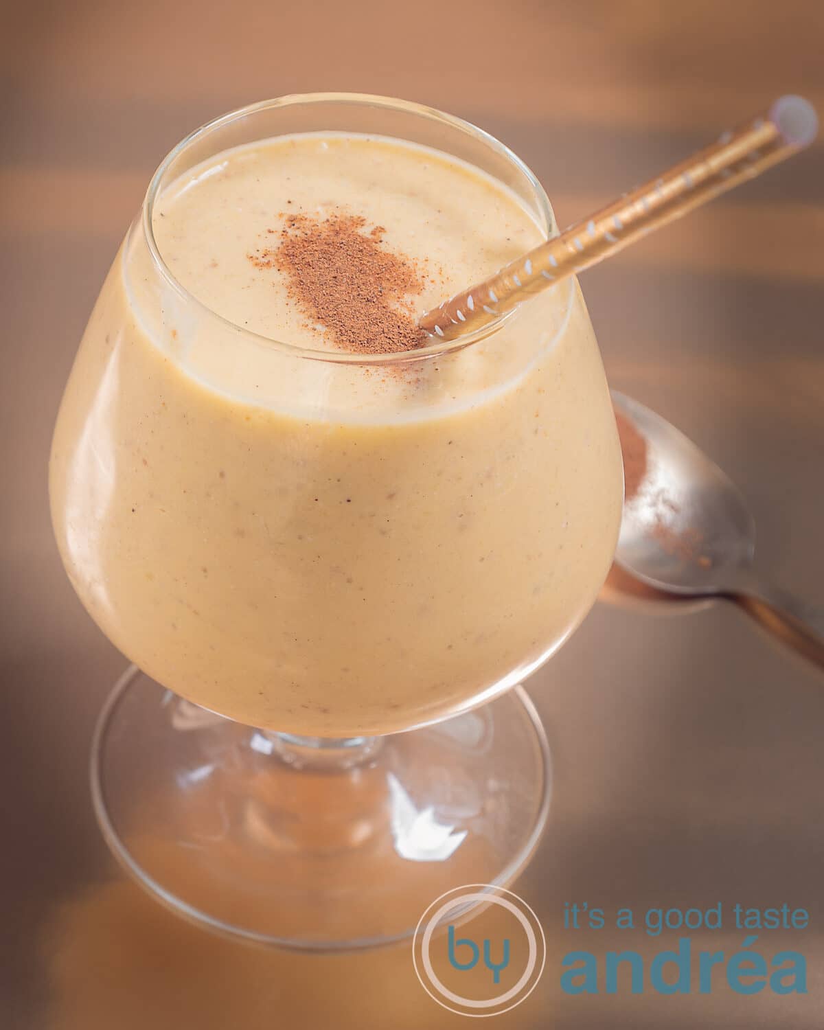 Een foto van een glas met een tropische mango-kokos-smoothie. Wat kaneel bovenop en een gouden rietje en achtergrond.