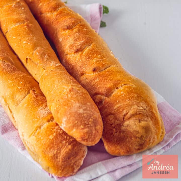 Een vierkante foto met drie zelfgebakken stokbroden in een stapeltje op een theedoek.