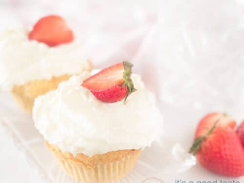 Een vierkante foto van twee cupcakes op een witte ondergrond op een schaaltje met een toef slagroom en een aardbei op top