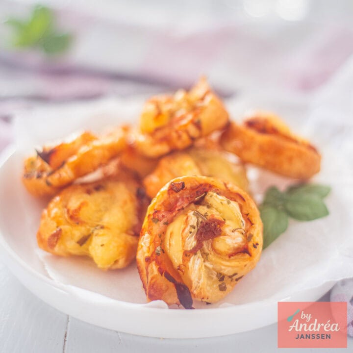 Een vierkante foto met een wit bord vol met bladerdeeghapjes met kaas en bacon.