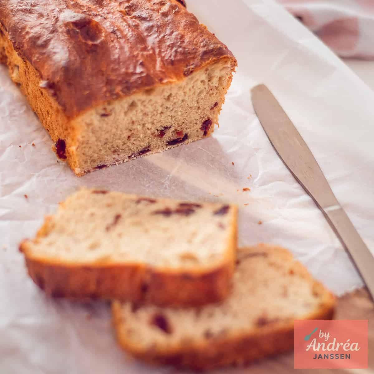 Een vierkante afbeelding van een cranberrybrood op bakpapier. Twee plakken zijn eraf gesneden.
