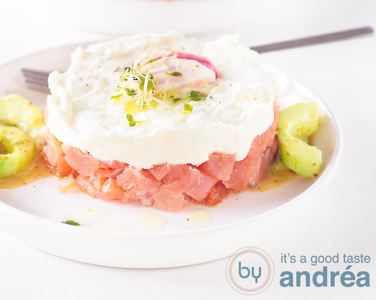 Een breedtefoto met een torentje van gerookte zalm, geitenkaas mousse op een bedje van komkommer tegen een witte ondergrond.