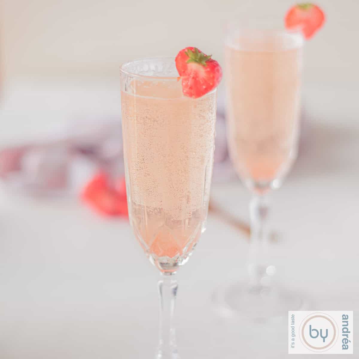 Een vierkante foto met twee champagne flutes gevuld met valentijnscocktail met een aardbeien garnering.